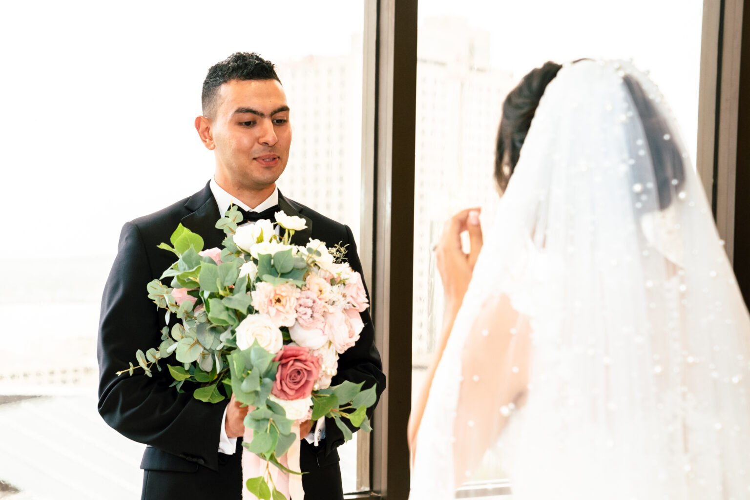 Groom holding a bouquet as he sees his veiled bride beside a bright window.