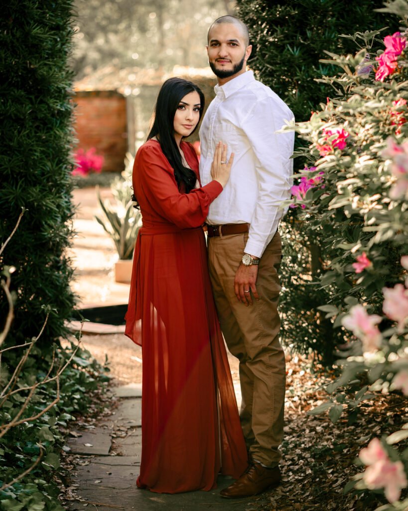 Couple in a red dress and white shirt surrounded by garden flowers.